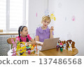 A mother and daughter wearing bunny ears are painting Easter eggs and looking at a laptop, celebrating the holiday together at home 137486568