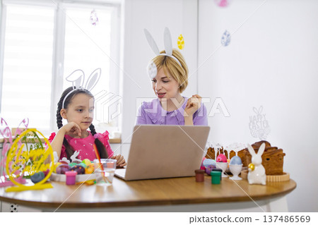 A mother and daughter wearing bunny ears participate in an online Easter egg decorating lesson on their laptop A mother and daughter wearing bunny ears participate in an online Easter egg decorating lesson on their laptop 137486569