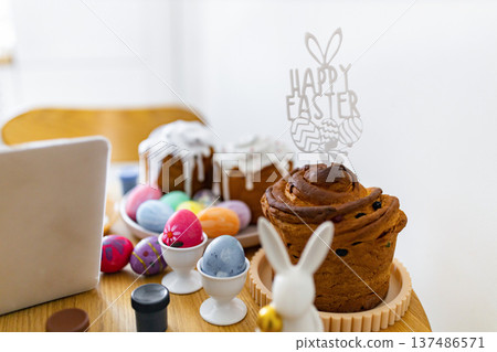 A festive table setting features traditional Easter cakes, colorful painted eggs in holders, and a 'Happy Easter' cake topper with bunny ears 137486571