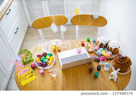 A wooden table is set up for Easter egg decorating with painted eggs, cakes, paints, and a laptop for inspiration or instructions A wooden table is set up for Easter egg decorating with painted eggs, cakes, paints, and a laptop for inspiration or instructions 137486574