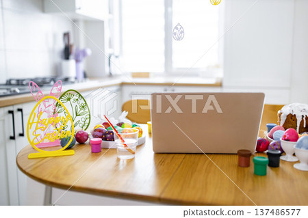 A table is set for Easter egg decorating with painted eggs, paints, brushes, and a laptop, suggesting a virtual celebration or tutorial A table is set for Easter egg decorating with painted eggs, paints, brushes, and a laptop, suggesting a virtual celebration or tutorial 137486577