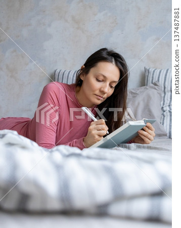 woman lies in bed writes in diary, writes emotions, wish list notebook. female writer plans workday. 137487881