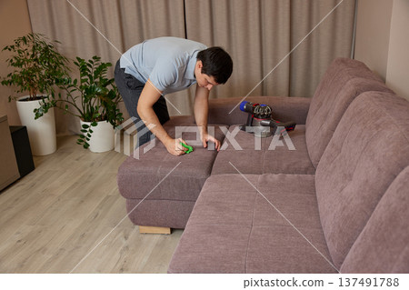 Man cleaning sofa upholstery at home using a powerful handheld vacuum cleaner Man cleaning sofa upholstery at home using a powerful handheld vacuum cleaner 137491788