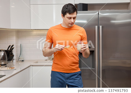 Young man standing in a modern kitchen, eating a sandwich and drinking milk 137491793