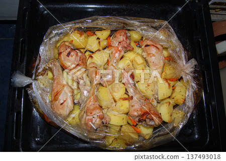 Roasted chicken drumsticks and potatoes baked in a bag. Chicken drumsticks and potatoes are roasted in a baking bag, placed on a black oven tray. 137493018