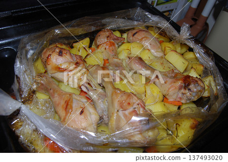Roasted chicken drumsticks and potatoes baked in a bag. Chicken drumsticks and potatoes are roasted in a baking bag, placed on a black oven tray. Roasted chicken drumsticks and potatoes baked in a bag. Chicken drumsticks and potatoes are roasted in a baking bag, placed on a black oven tray. 137493020