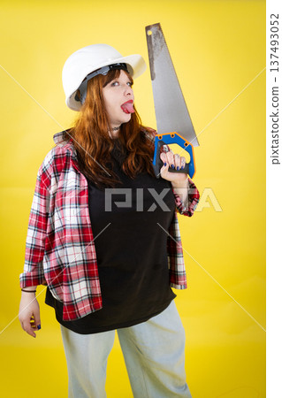 Woman wearing hard hat holding saw making funny face Woman wearing hard hat holding saw making funny face 137493052