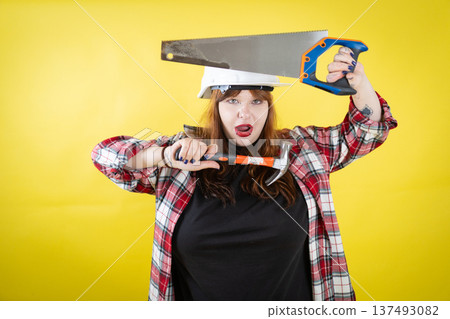 Frustrated woman holding hammer with saw on hard hat Frustrated woman holding hammer with saw on hard hat 137493082