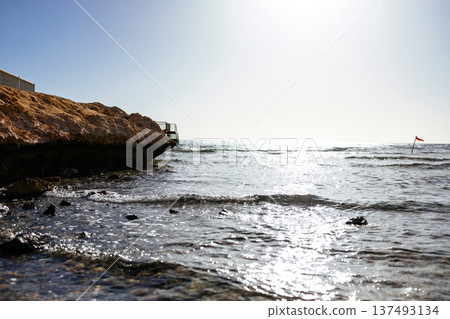Low angle view of a rocky sea coast with gentle waves washing over shallow water under bright sunlight. Sparkling rays of the sun reflected in the water. Red sea, Egypt. 137493134
