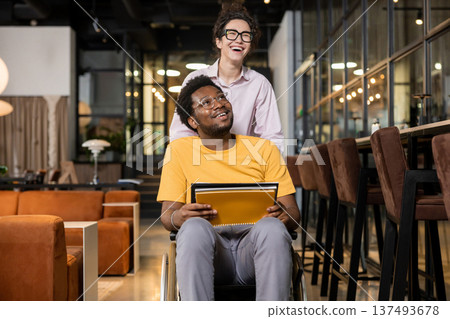 Woman helping disabled coworker by pushing his wheelchair in coworking interior 137493678