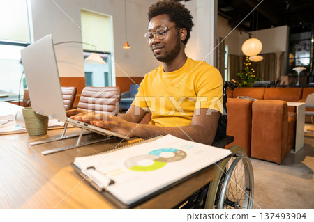 Man in wheelchair working on laptop with online business data in coworking space Man in wheelchair working on laptop with online business data in coworking space 137493904