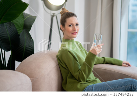 Pretty young woman in green sitting on the sofa with a glass of water 137494252