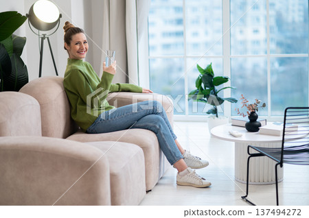 Pretty young woman in green sitting on the sofa with a glass of water 137494272