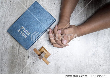 An overhead shot of a person's hands folded in prayer next to a blue Holy Bible and a small wooden cross on a textured grey surface. High angle view representing faith and devotion 137495330