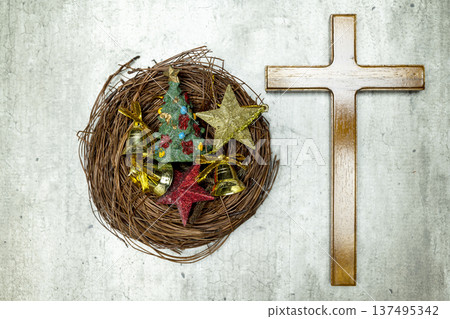 Top view of a wooden cross next to a bird's nest filled with Christmas tree ornaments, stars, and bells on a textured gray background 137495342