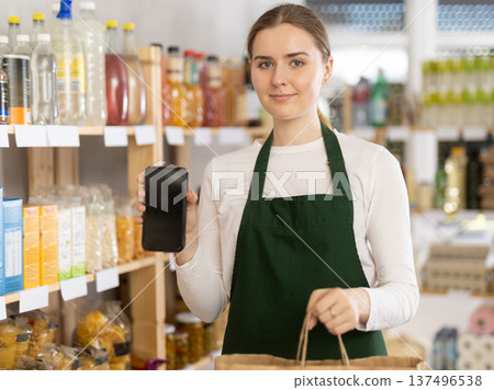 Girl seller in shop hold out hand with phone, points at screen with finger 137496538