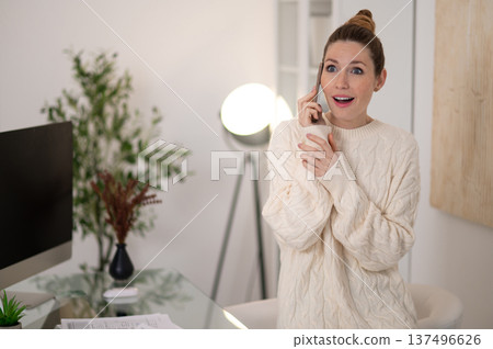 Woman in white sweater talking on the phone and looking surprised 137496626