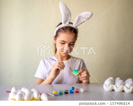 Young girl wearing bunny ears concentrates as she paints colorful Easter eggs at a spring craft table 137496814