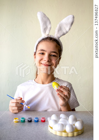Smiling girl wearing bunny ears painting a yellow Easter egg at table, feeling playful and creative Smiling girl wearing bunny ears painting a yellow Easter egg at table, feeling playful and creative 137496827