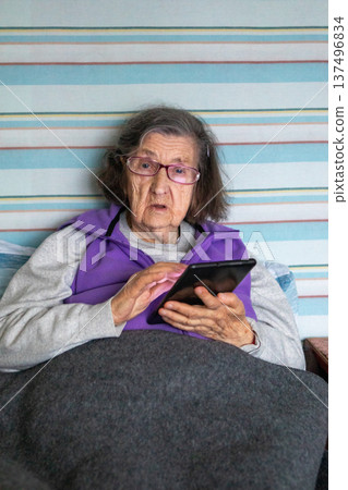 Elderly woman in bed using a tablet at home, wearing glasses and a purple vest Elderly woman in bed using a tablet at home, wearing glasses and a purple vest 137496834