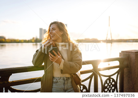 Young woman enjoys a sunny day by the river, using her smartphone while smiling in the warm sunlight 137497036