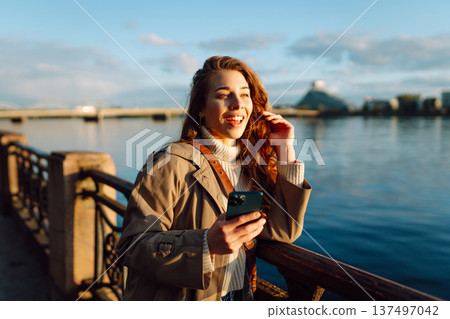 Young woman enjoys a sunny day by the river, using her smartphone while smiling in the warm sunlight Young woman enjoys a sunny day by the river, using her smartphone while smiling in the warm sunlight 137497042
