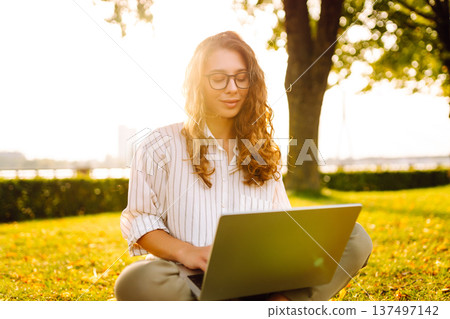 Young Woman sitting on grass ground, working on laptop. Freelance work, technology concept. Young Woman sitting on grass ground, working on laptop. Freelance work, technology concept. 137497142
