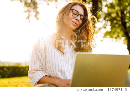 Young Woman sitting on grass ground, working on laptop. Freelance work, technology concept. Young Woman sitting on grass ground, working on laptop. Freelance work, technology concept. 137497143