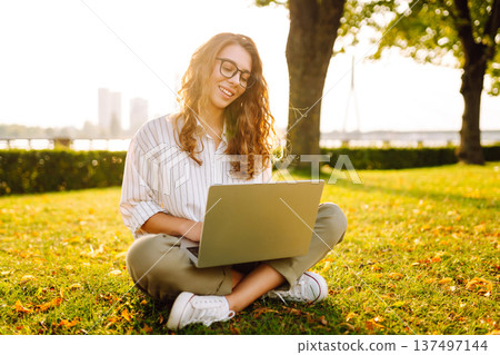 Young Woman sitting on grass ground, working on laptop. Freelance work, technology concept. Young Woman sitting on grass ground, working on laptop. Freelance work, technology concept. 137497144