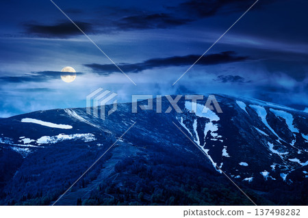 mysterious nature image of ridge with snow capped tops in spring landscape. mountain background wallpaper at night. full moon over greenery scenery with forested slopes under sky with clouds 137498282
