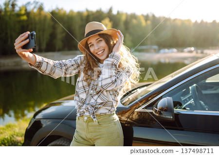 Happy woman on vacation taking selfie photo with smartphone near car. Stylish model on a car trip. Happy woman on vacation taking selfie photo with smartphone near car. Stylish model on a car trip. 137498711