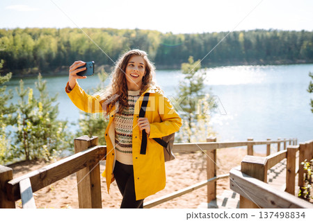 Happy woman with phone in hands taking selfie in forest. Young tourist enjoying spring day near lake 137498834