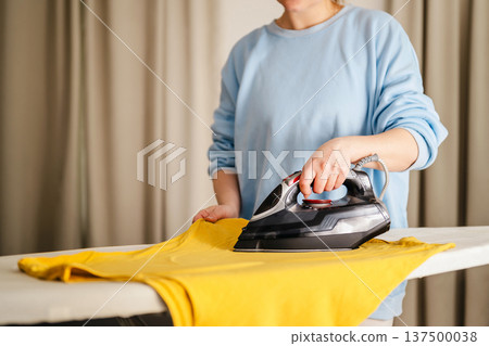 Woman in blue jamper, ironing t-shirts at home. Daily chores. Laundry day. Copy space 137500038