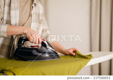 Home appliances. Woman ironing a green t-shirts at home. Daily chores. Laundry day. Neutral background with copy space Home appliances. Woman ironing a green t-shirts at home. Daily chores. Laundry day. Neutral background with copy space 137500053