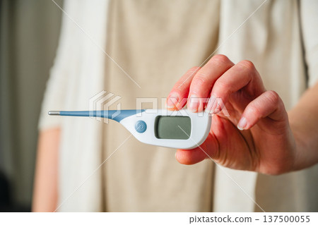 Electronic thermometer in female hands. Woman holding a digital thermometer. Flu and cold concept. Seasonal viruses Electronic thermometer in female hands. Woman holding a digital thermometer. Flu and cold concept. Seasonal viruses 137500055