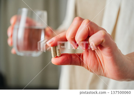 Woman holding two capsules pills in one hand and a glass with water, in other. Prescription medication, treatment sickness, seasonal cold flu concept 137500060