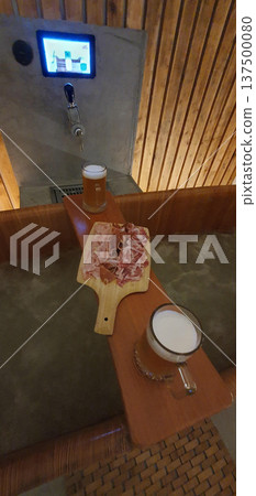 Phone photo of a wooden bath in a beer spa with jugs of beer and a charcuterie board. Relaxation and wellness scene mixing bathing ritual, food, and social leisure. 137500080