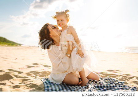 A mother and daughter enjoy a sunny beach stroll, as waves gently lap at their feet during sunset 137500138