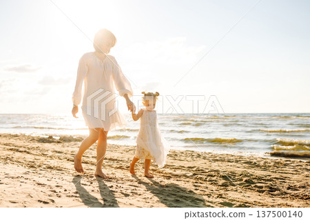 A mother and daughter enjoy a sunny beach stroll, as waves gently lap at their feet during sunset 137500140