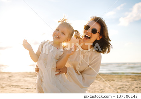 A mother and daughter enjoy a sunny beach stroll, as waves gently lap at their feet during sunset A mother and daughter enjoy a sunny beach stroll, as waves gently lap at their feet during sunset 137500142
