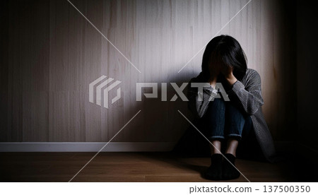 A young woman sits on the floor in a dark room with her back to the wall, her face buried in her knees. Deep sadness and despair. 137500350