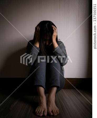 A young woman sits on the floor in a dark room with her back to the wall, her face buried in her knees. Deep sadness and despair. 137500351