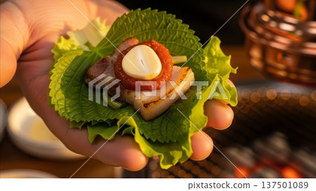 Korean BBQ pork belly lettuce wrap with gochujang sauce and garlic being held over grilled meat background 137501089