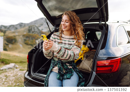 Young traveler woman sits on the rear of her car, smiling and using phone in scenic mountainous area Young traveler woman sits on the rear of her car, smiling and using phone in scenic mountainous area 137501392