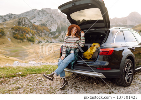 A traveler woman with curly hair and a hat smiles as she relaxes at the back of her black SUV 137501425