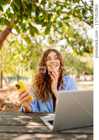 Happy woman with laptop and phone at table in an outdoor near lake. Business, blogging, freelancing. Happy woman with laptop and phone at table in an outdoor near lake. Business, blogging, freelancing. 137501869