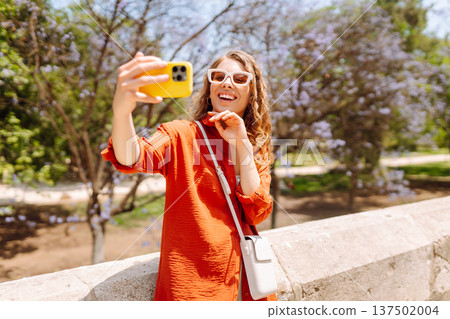 Young woman taking a cheerful selfie in blooming park on a sunny day. Joy concept. Active lifestyle. Young woman taking a cheerful selfie in blooming park on a sunny day. Joy concept. Active lifestyle. 137502004