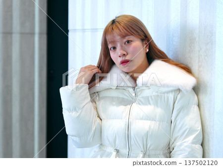 A female model leaning against a white wall. A sophisticated winter indoor portrait with a clean white outerwear. A female model leaning against a white wall. A sophisticated winter indoor portrait with a clean white outerwear. 137502120