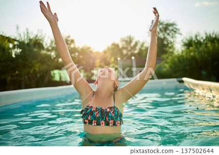 Teen girl enjoying a summer day in a pool, celebrating with arms raised under the warm sun 137502644