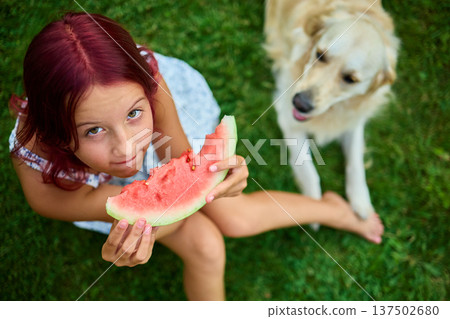 Teen girl enjoying watermelon while sitting on grass with a dog nearby during a sunny day in the backyard 137502680